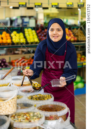 Asian female seller in hijab carefully puts large pickled olives in disposable lunch box Asian female seller in hijab carefully puts large pickled olives in disposable lunch box 136453752