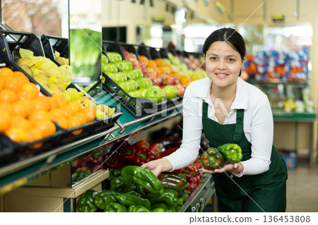 Asian female seller in apron chooses ripe bell pepper 136453808