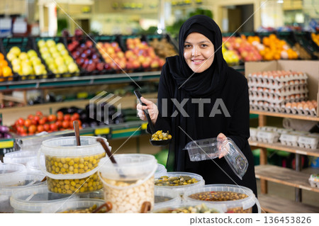 Asian woman in dark blue hijab puts handful of olives in disposable container with ladle Asian woman in dark blue hijab puts handful of olives in disposable container with ladle 136453826