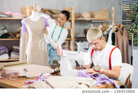 Young guy tailor sewing textile using sewing machine and asian female assistant working with fabric and mannequin in fashionable sewing atelier 136453868