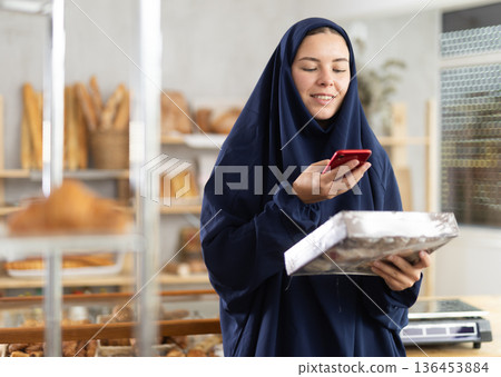 Woman in Muslim clothing scanning QR code on pastry box in bakery Woman in Muslim clothing scanning QR code on pastry box in bakery 136453884