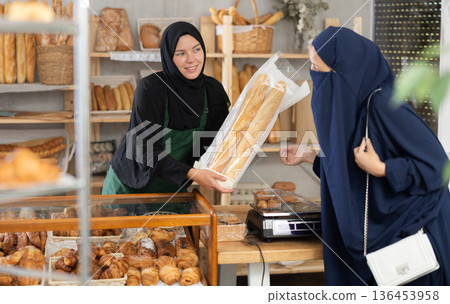 Muslim saleswoman offering baguettes to female customer in bakery Muslim saleswoman offering baguettes to female customer in bakery 136453958