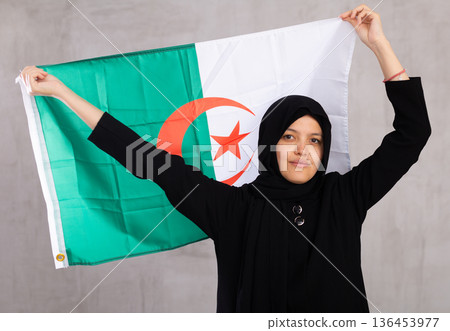 Young Muslim woman in black hijab holds unfurled flag of Algeria. Young Muslim woman in black hijab holds unfurled flag of Algeria. 136453977
