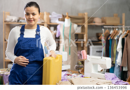 Experienced young repairwoman master posing with box of sewing machine repair tools near work table in fashionable sewing atelier 136453996