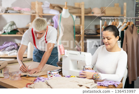 Attentive woman tailor sewing textile using sewing machine and young male assistant drawing pattern on sheet of paper using curves in fashionable sewing atelier 136453997