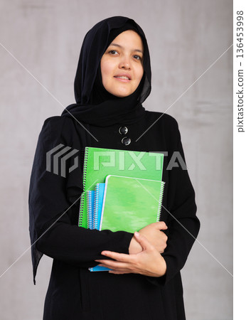 Portrait of young woman in hijab holding stack of notebooks 136453998