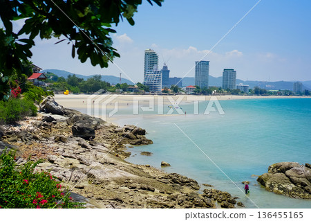 The landscape picture of the tranquil beach and azure sea at Hua Hin, Thailand with skyscraper view on the horizon line and blue sky. Beach rocks and tropical plants. The landscape picture of the tranquil beach and azure sea at Hua Hin, Thailand with skyscraper view on the horizon line and blue sky. Beach rocks and tropical plants. 136455165