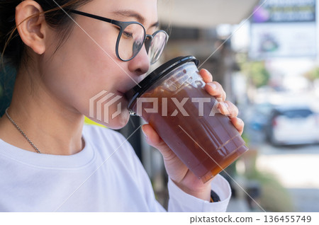Asian woman while drinking an iced Americano coffee. An iced americano is a refreshing coffee drink made with espresso and cold water, served over ice. Asian woman while drinking an iced Americano coffee. An iced americano is a refreshing coffee drink made with espresso and cold water, served over ice. 136455749