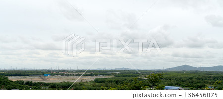 High-angle panoramic view of an industrial zone with factories on the horizon, surrounded by green forests, a dam reservoir, and mountains under a cloudy sky. Wide environmental landscape photo. 136456075