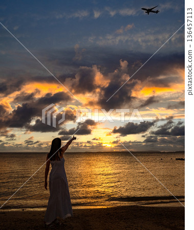 Image of a woman in a dress pointing at an airplane at sunset on a tropical beach Image of a woman in a dress pointing at an airplane at sunset on a tropical beach 136457113