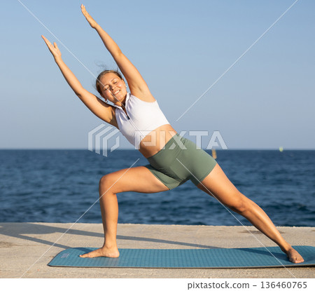 On windy day young slender woman does yoga near sea doing Utthita Parshvakonasana 136460765