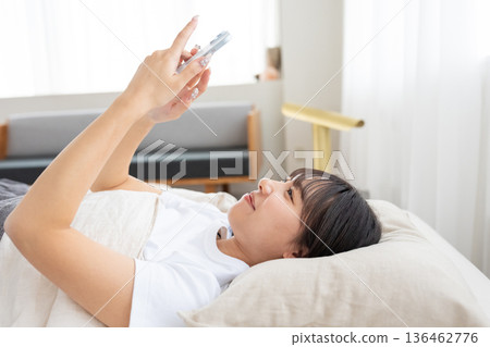 一位年輕的女士在床上操縱智能手機 一位年輕的女士在床上操縱智能手機 136462776