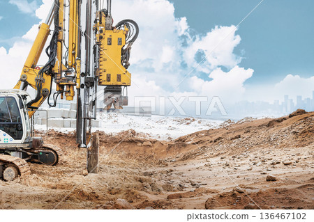 A drilling rig operates to install piles in the ground at a construction site during the day with a clear sky and distant buildings visible A drilling rig operates to install piles in the ground at a construction site during the day with a clear sky and distant buildings visible 136467102