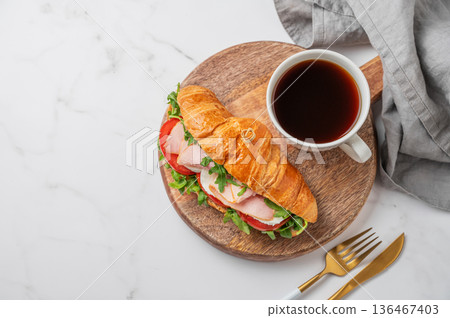 Croissant sandwich with ham, mozzarella cheese, tomato, arugula on a wooden board and cup of coffee Croissant sandwich with ham, mozzarella cheese, tomato, arugula on a wooden board and cup of coffee 136467403