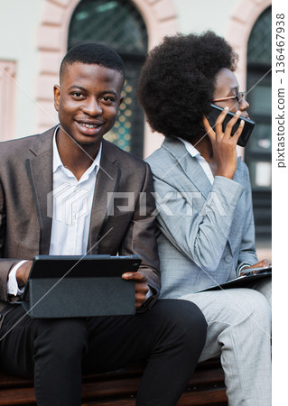 A man and a woman in business attire sit on a bench outdoors, one using a tablet and the other talking on a phone A man and a woman in business attire sit on a bench outdoors, one using a tablet and the other talking on a phone 136467938