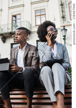 A man and a woman in business attire sit outdoors on a bench, with the woman talking on a mobile phone and the man holding a tablet A man and a woman in business attire sit outdoors on a bench, with the woman talking on a mobile phone and the man holding a tablet 136467940