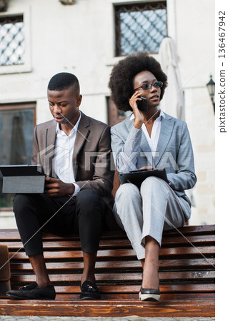 A man and a woman in business attire sit on a wooden bench outdoors, engaged with their digital devices and phones 136467942