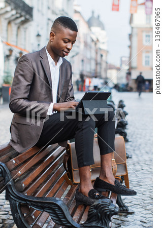 A stylish young man in a suit jacket and loafers sits on a park bench in a European city, engrossed in his tablet A stylish young man in a suit jacket and loafers sits on a park bench in a European city, engrossed in his tablet 136467965