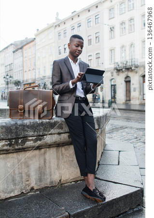 A young Black man in a suit jacket and trousers stands outdoors in a European city, looking at a tablet device while his leather briefcase rests beside him A young Black man in a suit jacket and trousers stands outdoors in a European city, looking at a tablet device while his leather briefcase rests beside him 136467968