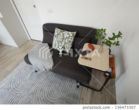 Compact black folding sofa bed with throw and pillows, and side table. Furniture for small living room. Light walls, gray carpet. Compact black folding sofa bed with throw and pillows, and side table. Furniture for small living room. Light walls, gray carpet. 136473307