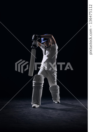 Youth cricket player raising bat in overhead pose on dark studio set. Youth cricket player raising bat in overhead pose on dark studio set. 136475112