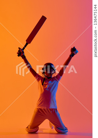 Celebrating young cricket player raising bat and arms in neon light. Celebrating young cricket player raising bat and arms in neon light. 136475144