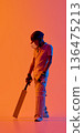 Young cricket batsman standing with bat grounded on vibrant orange gradient background. 136475213