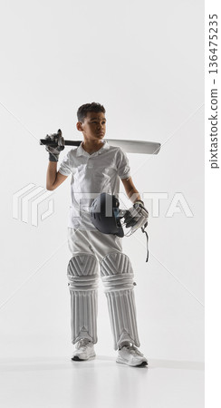 Young cricket player standing with bat on shoulder holding helmet in studio. 136475235