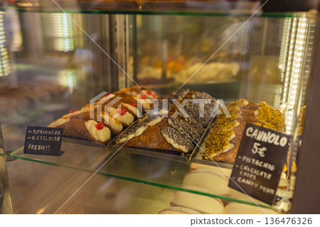 Traditional Italian cannoli pastries with various toppings displayed in a bakery glass showcase, featuring classic Sicilian desserts under warm interior lighting. Traditional Italian cannoli pastries with various toppings displayed in a bakery glass showcase, featuring classic Sicilian desserts under warm interior lighting. 136476326