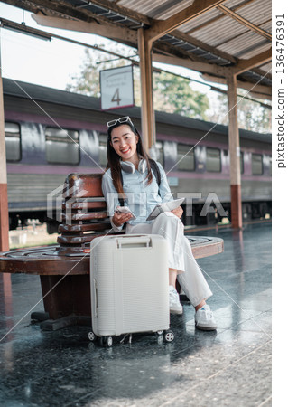 Smiling woman sitting at a train station with luggage and tablet, ready for a travel adventure. Smiling woman sitting at a train station with luggage and tablet, ready for a travel adventure. 136476391