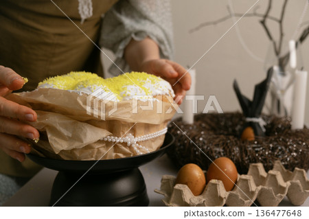 In home clothes, she arranges Easter cakes and colorful eggs on the table, preparing it with love and warmth. In home clothes, she arranges Easter cakes and colorful eggs on the table, preparing it with love and warmth. 136477648