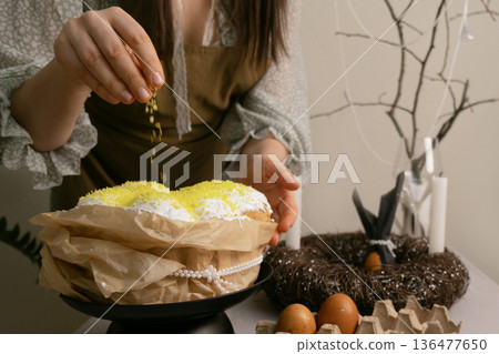 In final moments before Easter, woman carefully sprinkles coconut on Easter cake, creating a festive holiday atmosphere. 136477650