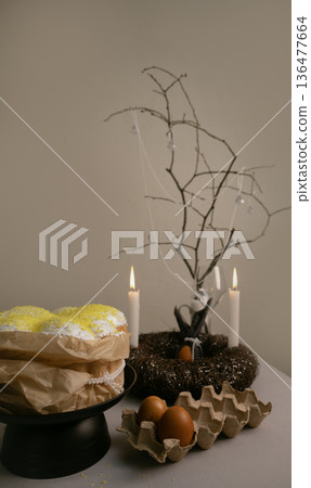 Cozy Easter composition with cake in white icing, colorful eggs in tray, candles, and twig wreath creating festive atmosphere. Cozy Easter composition with cake in white icing, colorful eggs in tray, candles, and twig wreath creating festive atmosphere. 136477664