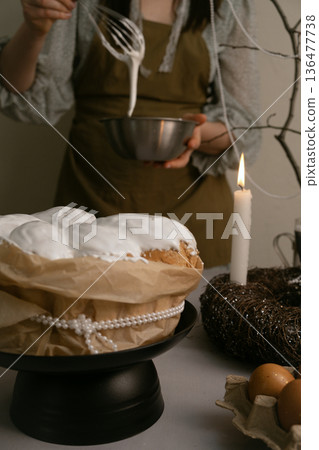 Cozy Easter moment in kitchen, she carefully spreads icing on cake, with eggs, candles, wreath on table, beaded branch in background. 136477738