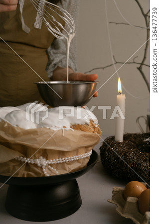 Woman brushes white icing on Easter cake, creating festive atmosphere with eggs, candles, wreath on table, beaded branch in background. Woman brushes white icing on Easter cake, creating festive atmosphere with eggs, candles, wreath on table, beaded branch in background. 136477739