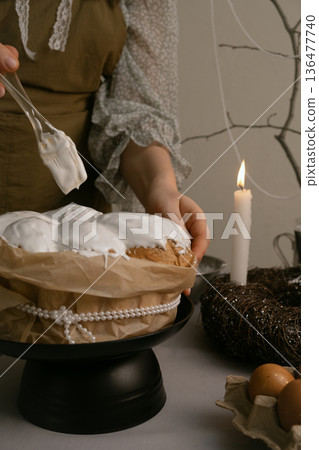 Easter preparation, she spreads white icing on cake, with eggs, candles, twig wreath on table, and decorative beaded branch in background. Easter preparation, she spreads white icing on cake, with eggs, candles, twig wreath on table, and decorative beaded branch in background. 136477740