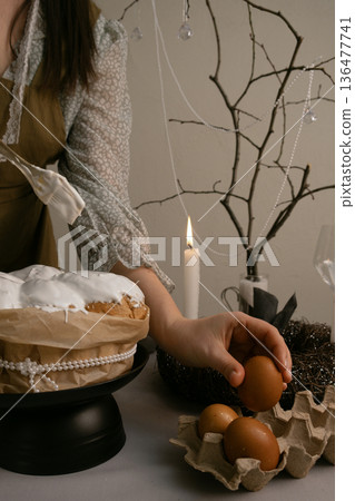 Woman in home clothes carefully decorates Easter cake with white icing, eggs, candles, wreath, and beaded branch creating festive atmosphere. Woman in home clothes carefully decorates Easter cake with white icing, eggs, candles, wreath, and beaded branch creating festive atmosphere. 136477741