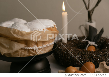Easter composition with cake in white icing, colorful eggs, and candles in a twig nest on a festive holiday table. Easter composition with cake in white icing, colorful eggs, and candles in a twig nest on a festive holiday table. 136477744