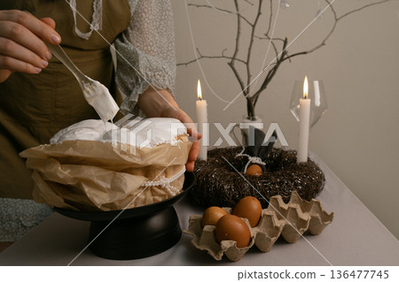 Woman uses brush to spread white icing on Easter cake, table decorated with eggs, candles, wreath, and beaded branch in background. Woman uses brush to spread white icing on Easter cake, table decorated with eggs, candles, wreath, and beaded branch in background. 136477745