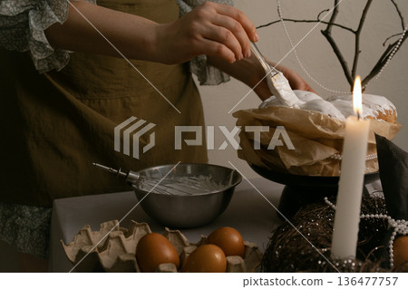 Preparing for Easter, she spreads white icing on cake, with eggs, candles, and decorative wreath on table, beaded branch in background. Preparing for Easter, she spreads white icing on cake, with eggs, candles, and decorative wreath on table, beaded branch in background. 136477757