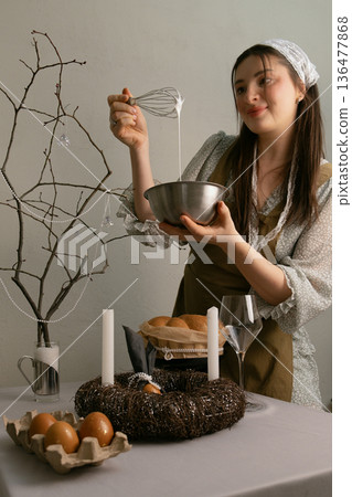 Woman in headscarf and apron whipping white cream for Easter cake at festive table 136477868