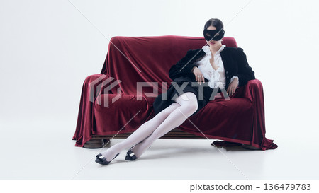 Woman with black mask reclining on red velvet sofa in white studio. 136479783