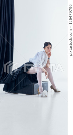 Female model sitting on black box in layered skirt and sheer tights in minimalist studio. 136479796