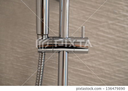 Chrome shower head holder with a flexible hose on a sliding rail, reflecting light against a textured bathroom wall for modern hygiene and personal care 136479890