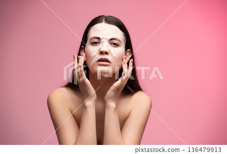 Young woman applying nourishing sheet mask to her face, having a skincare routine for beauty and wellness, doing self-care on a pink background Young woman applying nourishing sheet mask to her face, having a skincare routine for beauty and wellness, doing self-care on a pink background 136479913