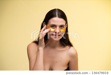 Young woman applying golden eye patches for under-eye skincare treatment, nurturing skin with beauty product, showing self-care routine against a yellow background 136479919