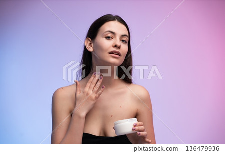 Young woman applying moisturizer cream on her neck while looking at the camera, highlighting a self-care beauty routine for hydration and healthy skin Young woman applying moisturizer cream on her neck while looking at the camera, highlighting a self-care beauty routine for hydration and healthy skin 136479936