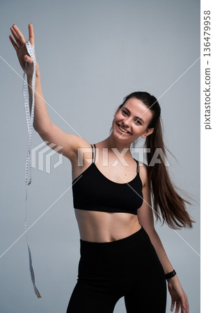 Happy young woman in sports attire smiling at the camera, showing a white measuring tape, symbolizing achievement, diet progress, and a healthy lifestyle 136479958