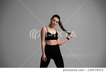 Woman posing in black sportswear, holding her ponytail in a studio, promoting fitness, healthy lifestyle, exercise, wellness, and active living Woman posing in black sportswear, holding her ponytail in a studio, promoting fitness, healthy lifestyle, exercise, wellness, and active living 136479972