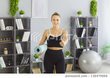 Smiling woman giving thumbs up while holding a yoga mat in a bright home gym space 136479989